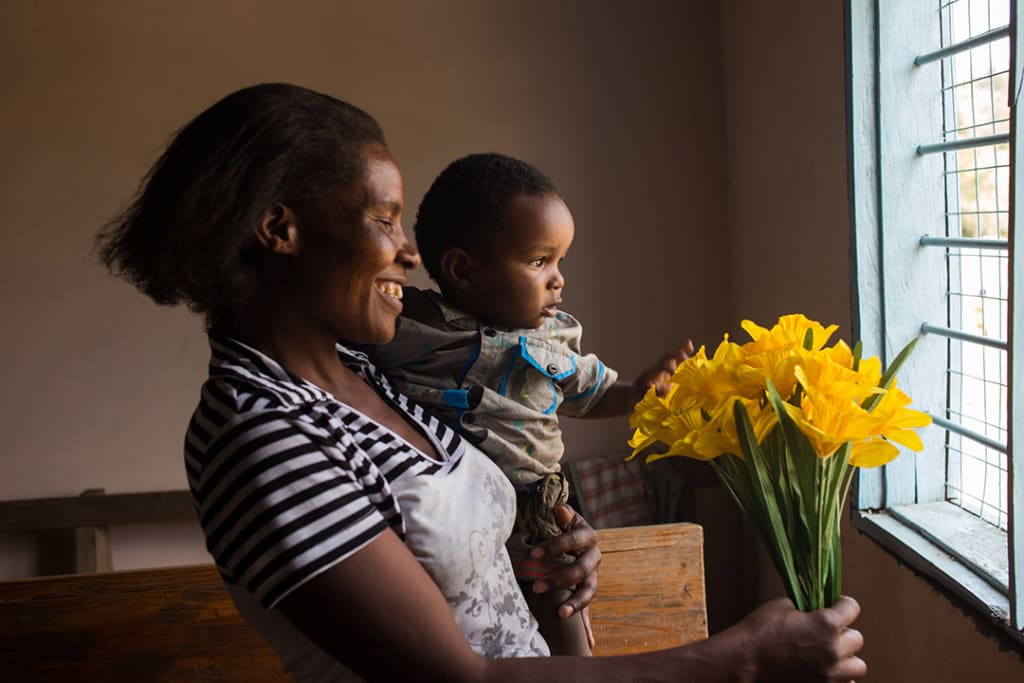 Selena, a Ugandan mother, holds her infant son Jemaal and bouquet of yellow flowers. They both look out a window.
